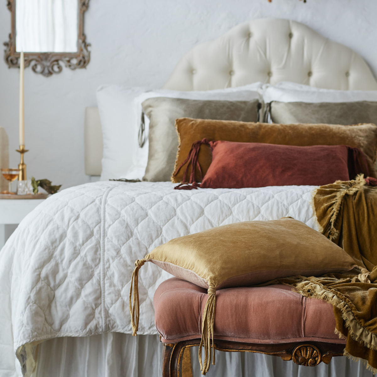 : Harlow coverlet in winter white on a neatly made bed against a grand tufted headboard. Warm brown and gold toned throw pillows and blankets in silk charmeuse and silk velvet provide contrast to the quilted cotton velvet - end of bed view.