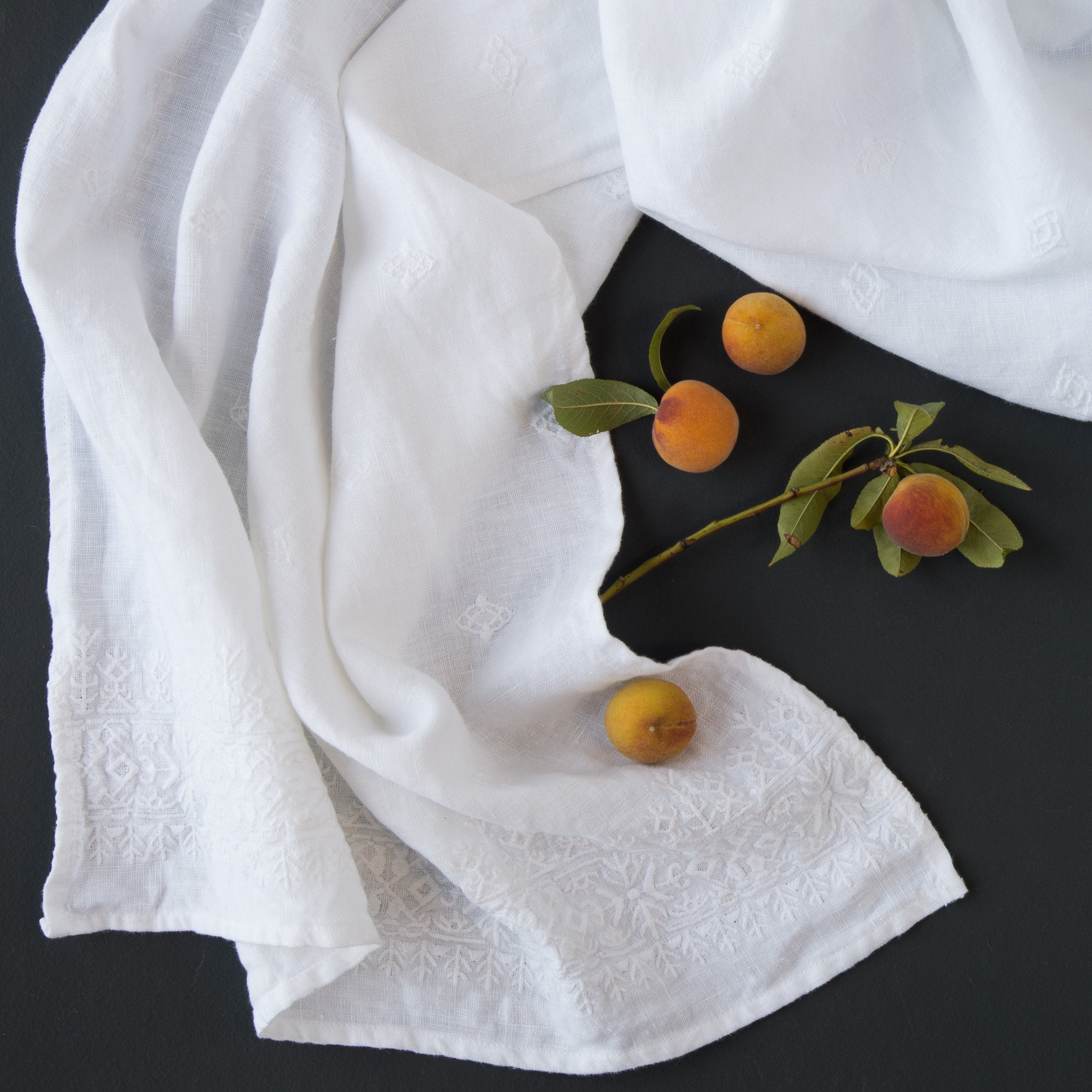 : White Ines guest towel draped on a dark background scattered with baby apricots and green leaves - overhead view.