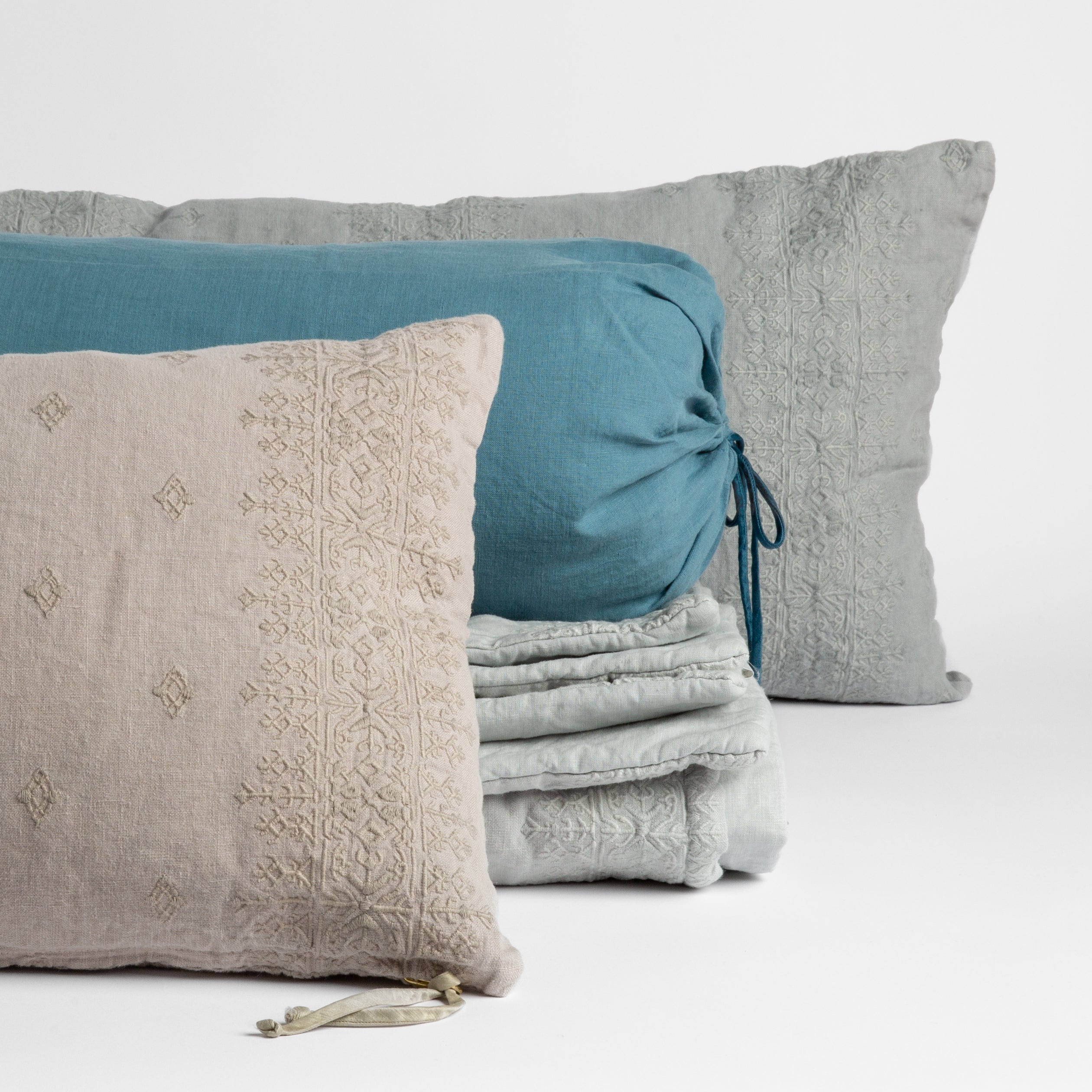 : embroidered midweight linen throw pillows and folded bed end sized throw blanket with linen bolster straight on against a white background.