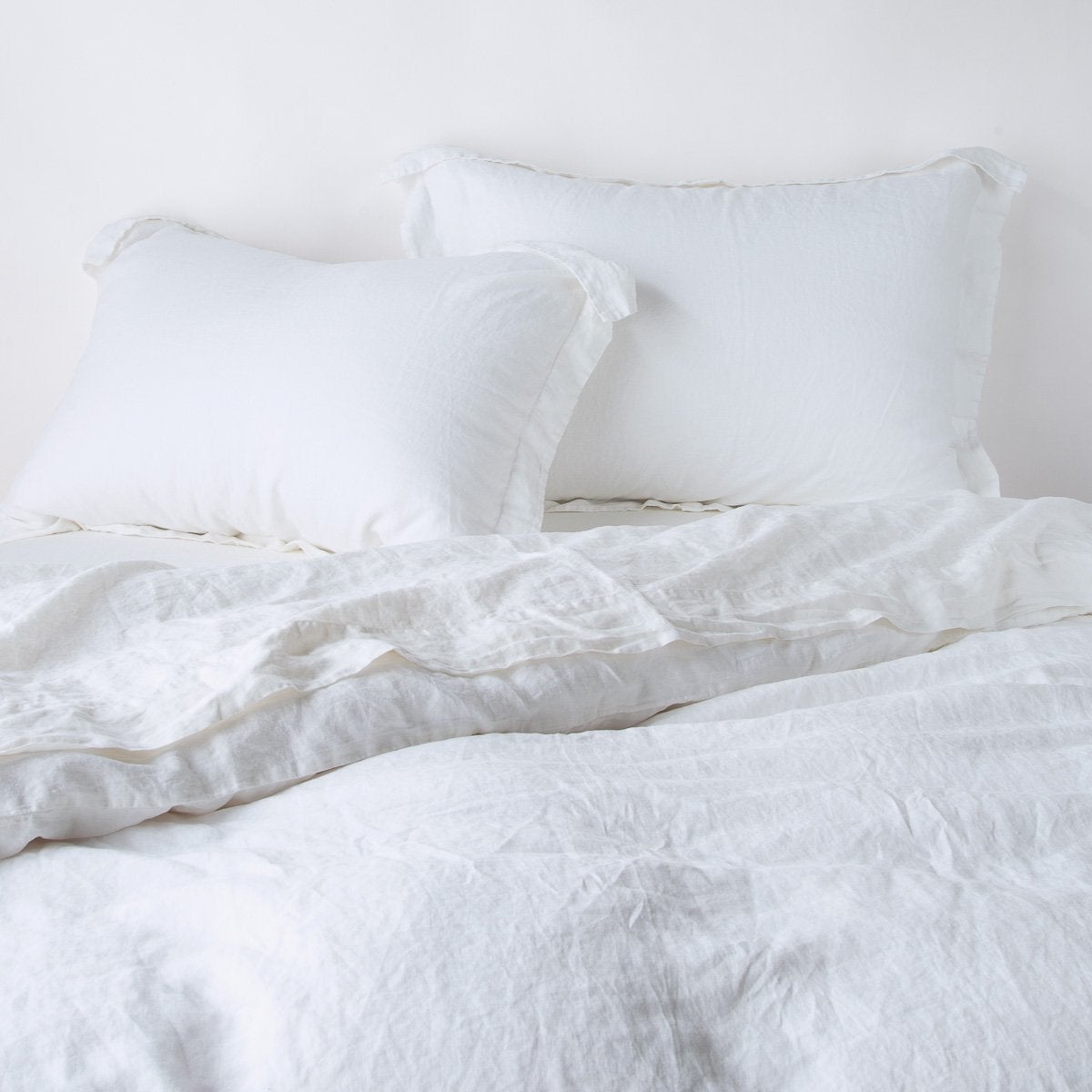 : Linen duvet cover in white, partially folded back on a bed with matching sheets and shams on a plain background - cropped end of bed view.