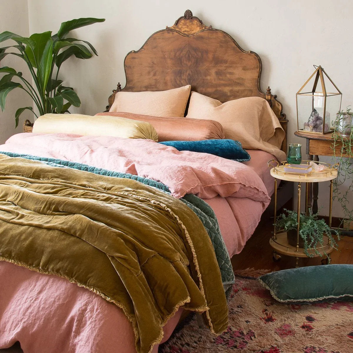 : Paloma bolsters and sleeping pillows layered with linen and silk velvet, in pink, gold, blue and green tones. Three-quarter angle showing dramatic wooden headboard and tall greenery.