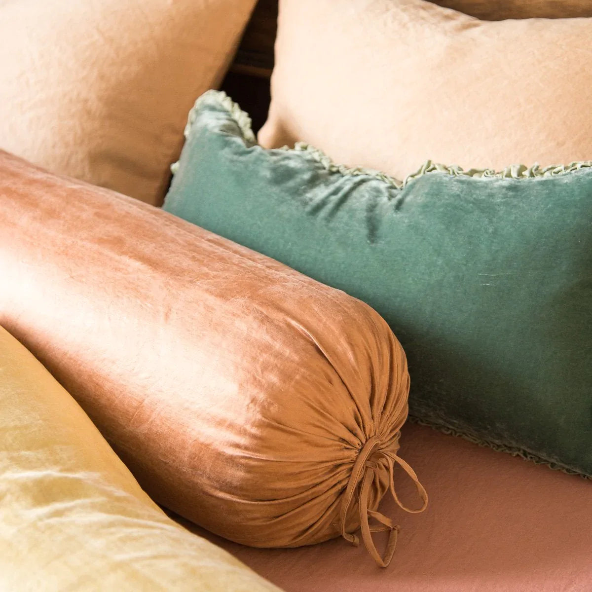 : Paloma bolsters and sleeping pillows layered with linen and silk velvet, in pink, gold, and green tones - close-up three-quarter angle.