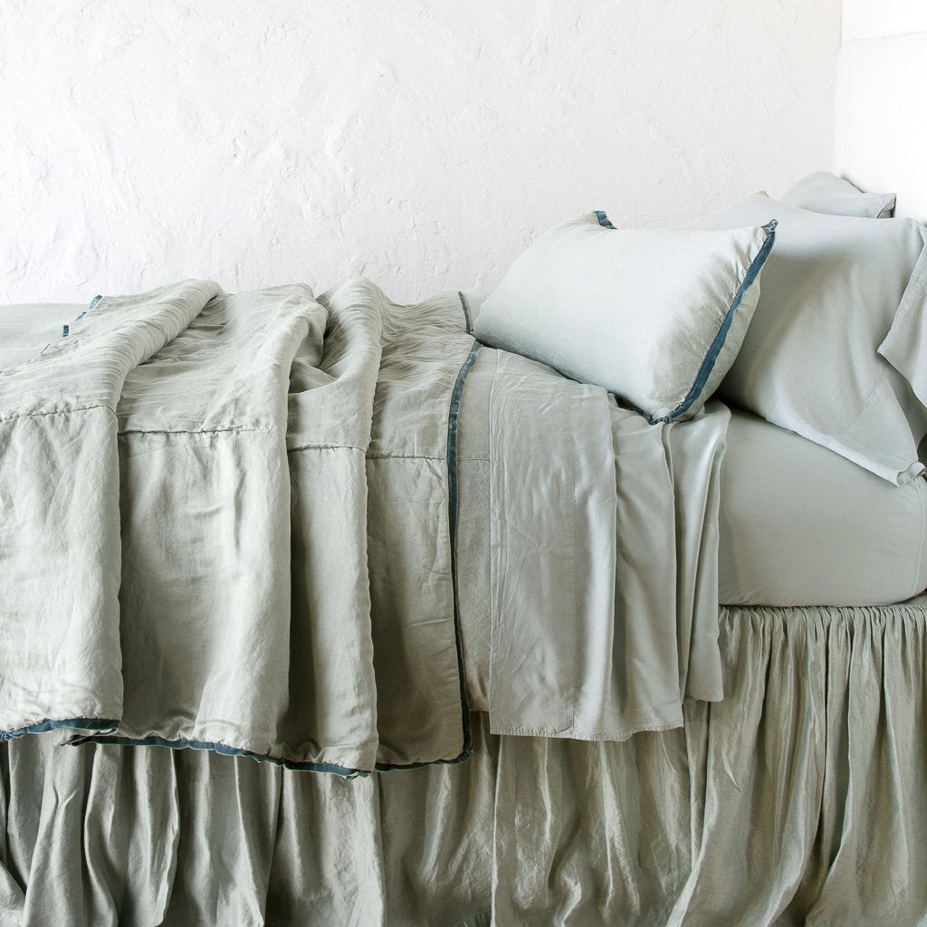 : Paloma throw blanket rumpled on a charmeuse monochromatic bed - eucalyptus, side view.