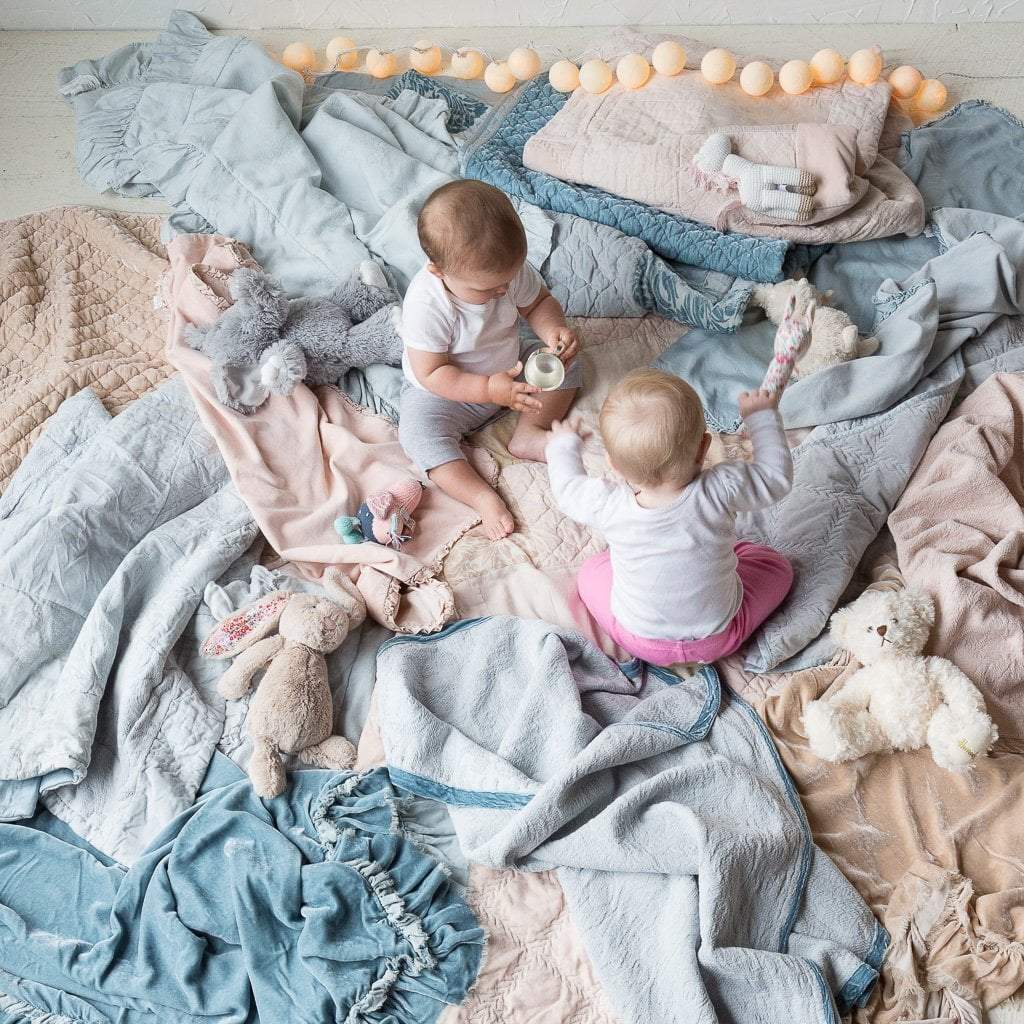 : An image taken from an overhead angle of two babies sitting and playing among a lot of rumpled blankets in cloud, sterling and pearl.