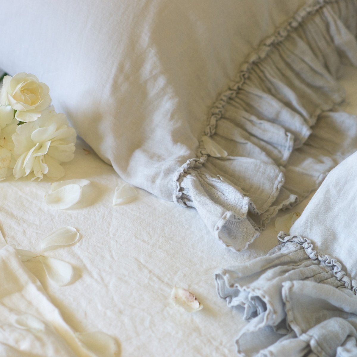 [allvariants]: Linen Whisper pillow cases in cloud, with light cream toned sheets and flower petals - close-up overhead angle showcases ruffle detail trim.