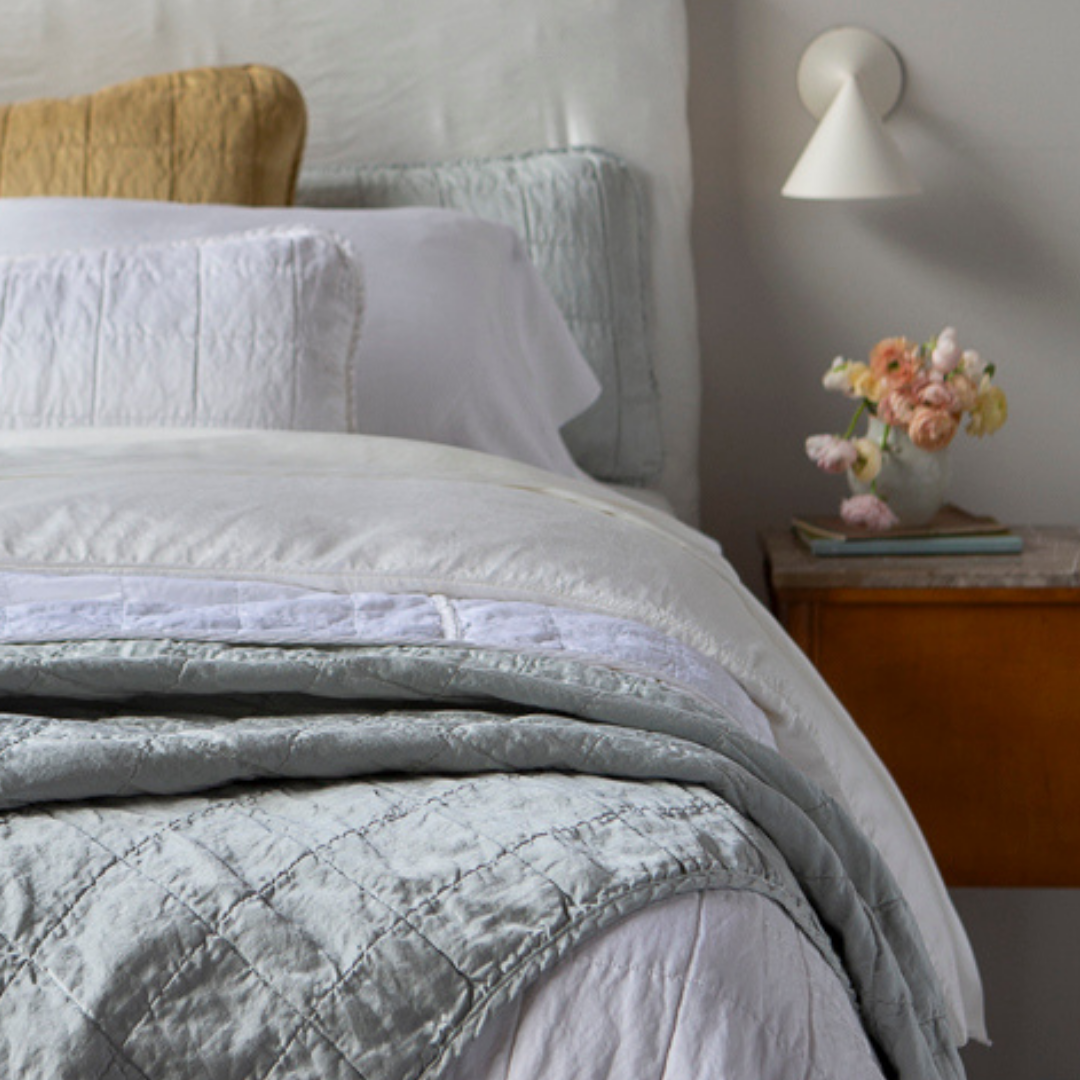 [allvariants]: Shot from the foot of the bed, a quilted charmeuse blanket is in the foreground and the pillows at the head of the bed are slightly out of focus. 