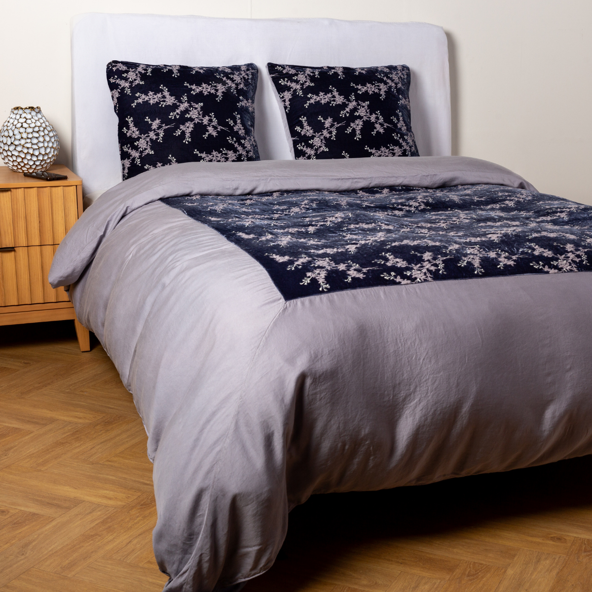 French Lavender: A ¾ angle shot of an embroidered silk velvet and tencel duvet cover and matching shams with a vase and cell phone on the side table. 