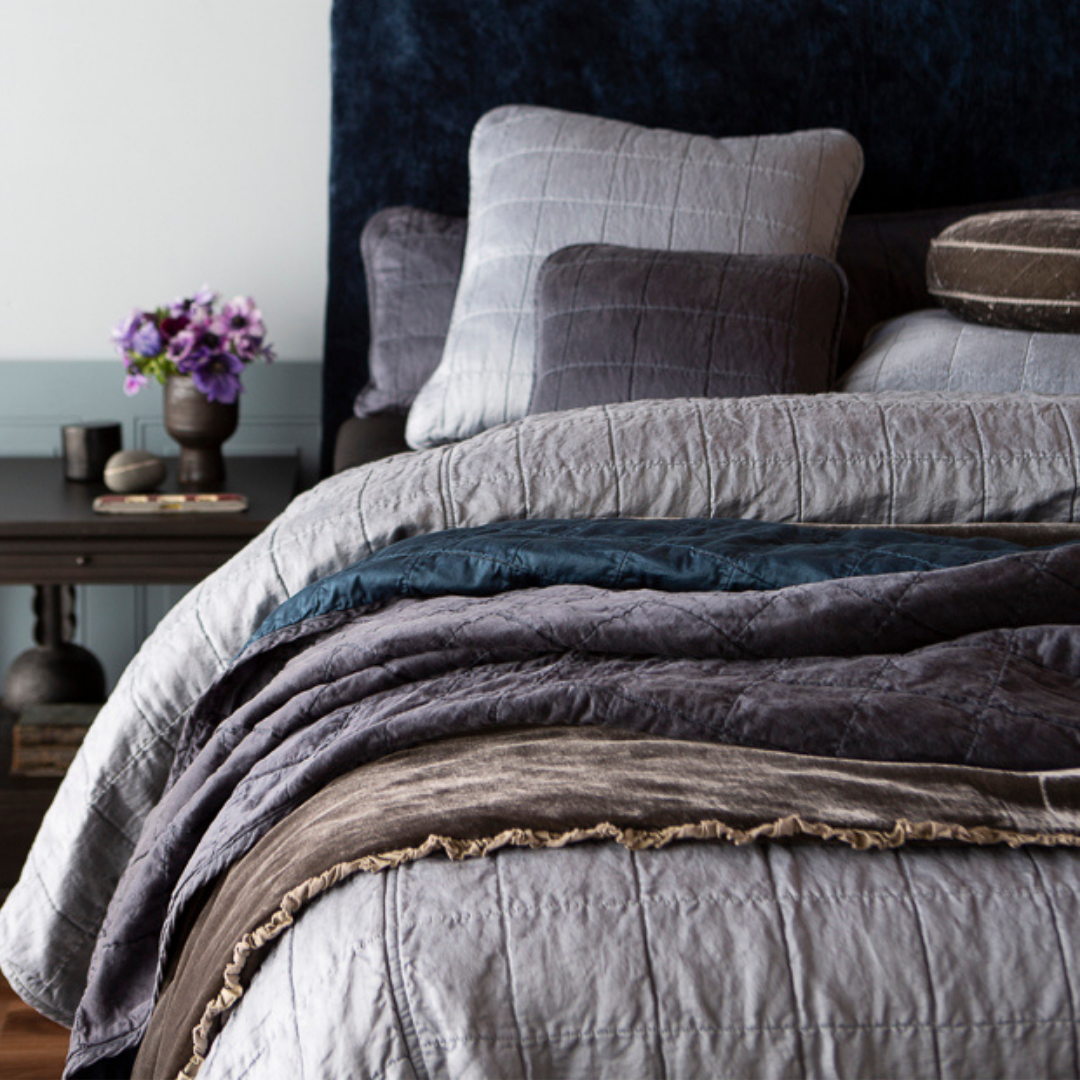 [allvariants]: a quilted charmeuse coverlet layered with a bed end blanket and throw blanket - shown from the foot of the bed, the pillows near the headboard are slightly out of focus as are the flowers on the side table.