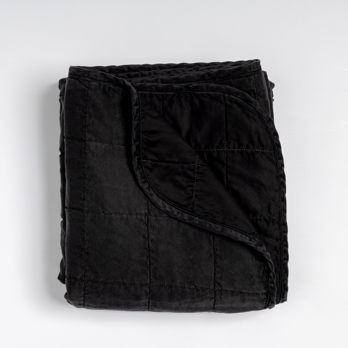Corvino: an overhead shot of a quilted charmeuse blanket in corvino, our classic black, with a corner folded down, showing its cotton sateen back. 