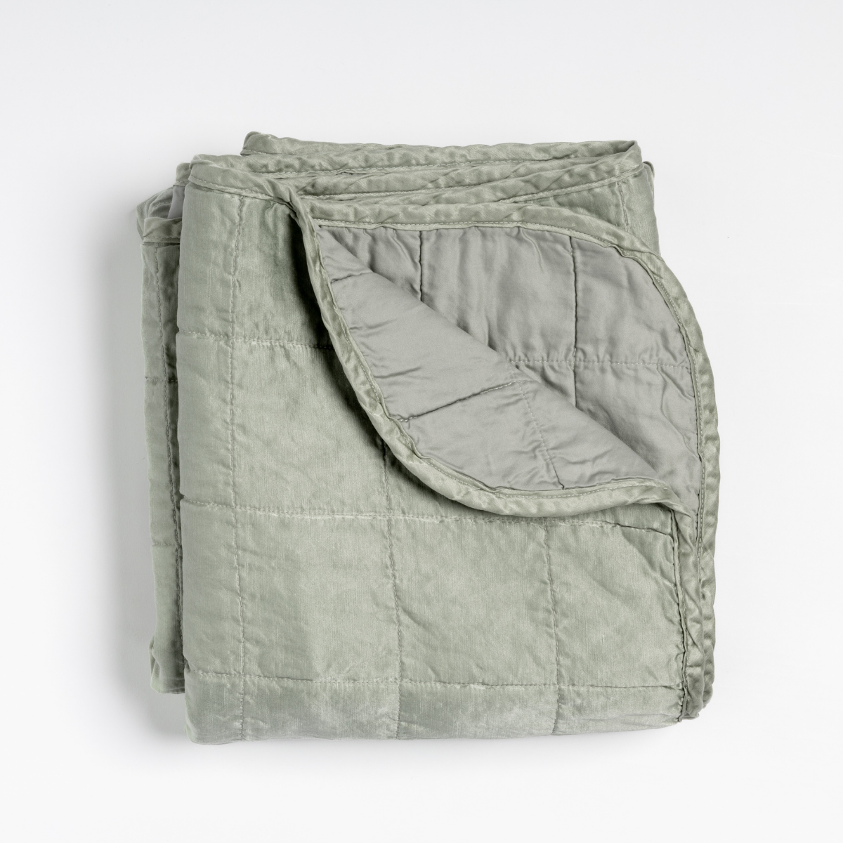 Eucalyptus: an overhead shot of a quilted charmeuse blanket in eucalyptus, a soft light green, with a corner folded down, showing its cotton sateen back. 