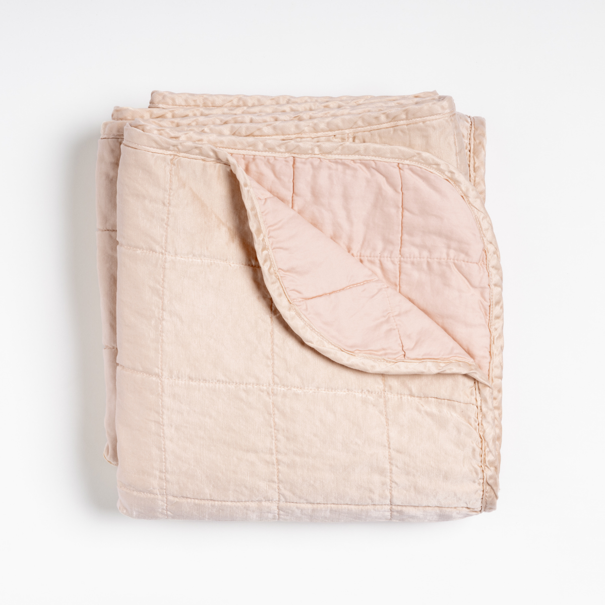 Pearl: an overhead shot of a quilted charmeuse blanket in pearl, a soft rose pink tone, with a corner folded down, showing its cotton sateen back. 
