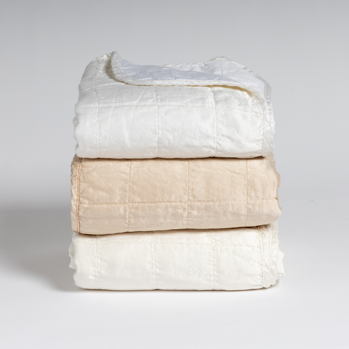 A stack of three square-quilted charmeuse blankets shot straight on against a white background. Colors from top to bottom are White, Pearl, and Winter White.