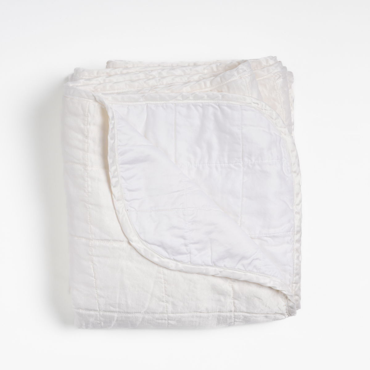 White: an overhead shot of a quilted charmeuse blanket in white, with a corner folded down, showing its cotton sateen back. 
