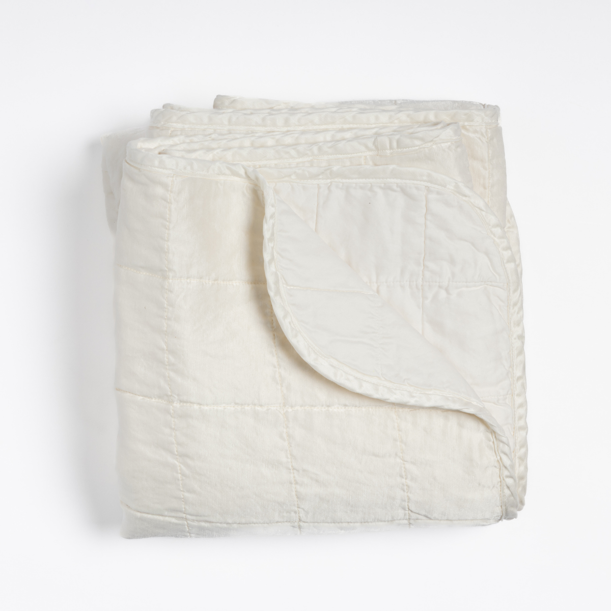Winter White: an overhead shot of a quilted charmeuse blanket in winter white, a softer and warmer tone than our classic white, with a corner folded down, showing its cotton sateen back. 