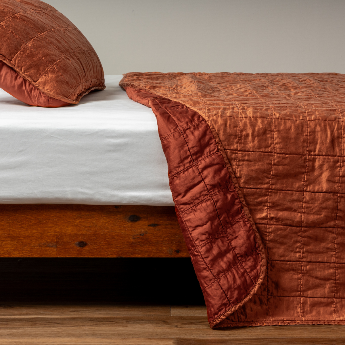 Autumn: A side shot of a square quilted charmeuse coverlet in autumn, a rich terracotta tone, with its matching sham, the corner of the coverlet peeled back, revealing the cotton sateen back. 