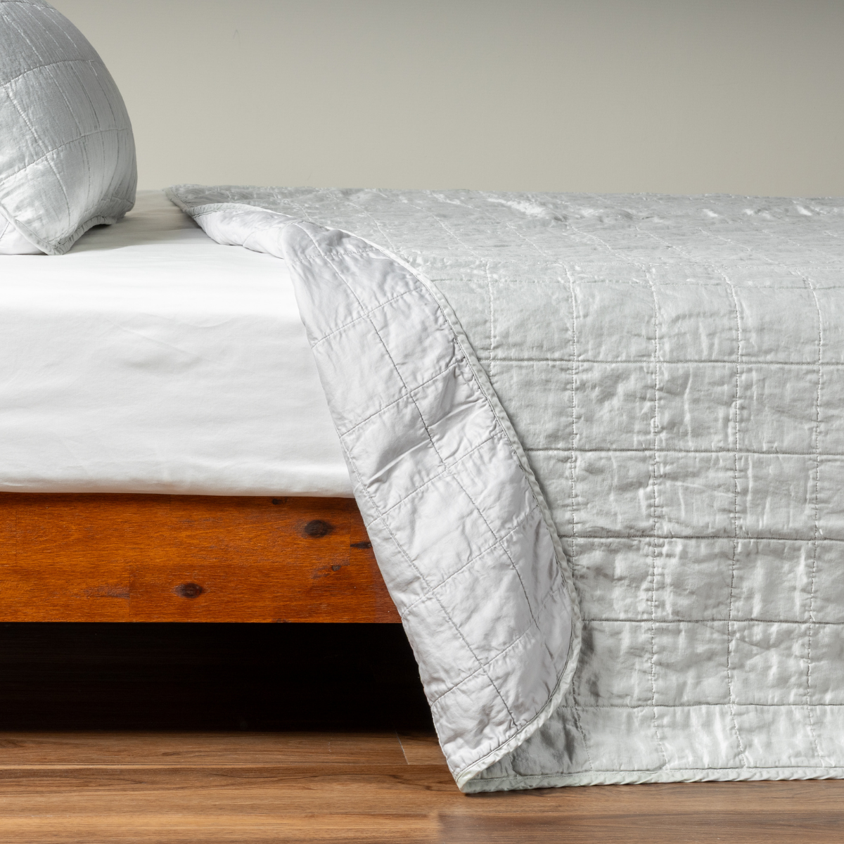 Cloud: A side shot of a square quilted charmeuse coverlet in cloud, a subtle sky blue-grey, with its matching sham, the corner of the coverlet peeled back, revealing the cotton sateen back. 