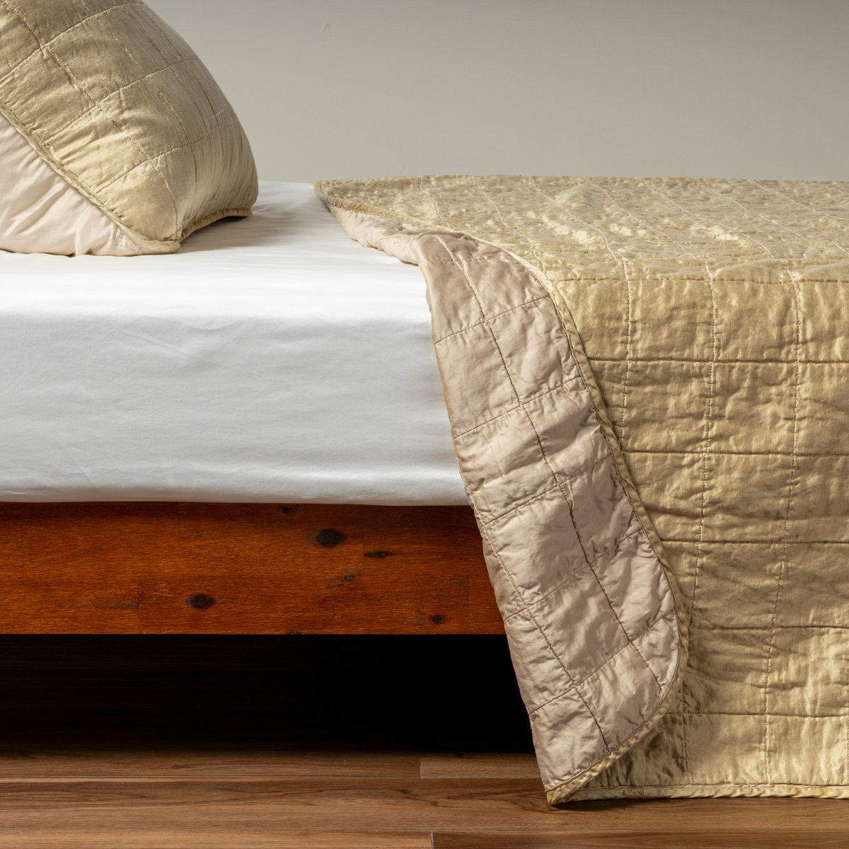 Honeycomb: A side shot of a square quilted charmeuse coverlet in honeycomb, a warm golden tone, with its matching sham, the corner of the coverlet peeled back, revealing the cotton sateen back. 