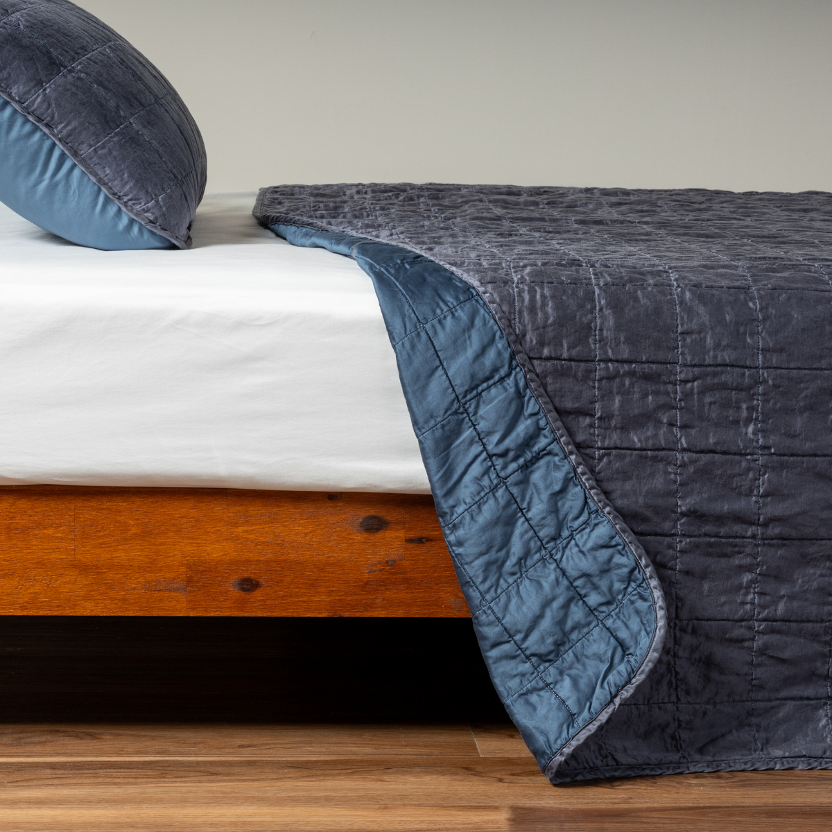 Midnight: A side shot of a square quilted charmeuse coverlet in midnight, a rich indigo tone, with its matching sham, the corner of the coverlet peeled back, revealing the cotton sateen back. 