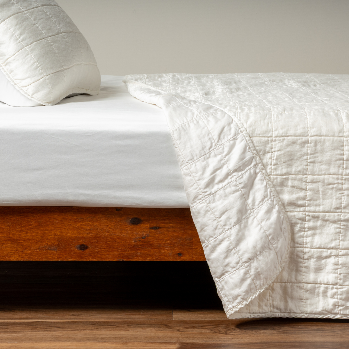 Winter White: A side shot of a square quilted charmeuse coverlet in winter white, a softer and warmer tone than our classic white, with its matching sham, the corner of the coverlet peeled back, revealing the cotton sateen back. 