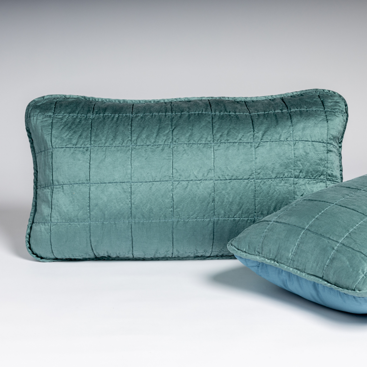 Cenote: A pair of shams shown against a white background in cenote, a vibrant ocean-inspired blue-green, with one sham upright and the other on its side to show the cotton sateen back and square-quilted charmeuse front. 