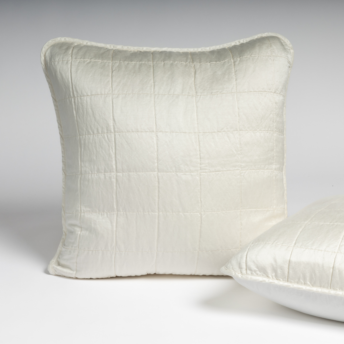 Winter White: A pair of shams shown in winter white, a softer and warmer tone than our classic white, against a white background with one sham upright and the other on its side to show the cotton sateen back and square-quilted charmeuse front. 