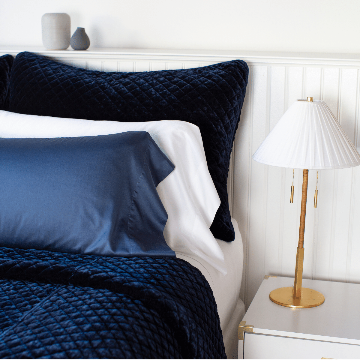 : Silk velvet quilted shams in deep midnight blue with matching coverlet, layered simply with white and midnight sleeping pillows - cropped three-quarter angle.