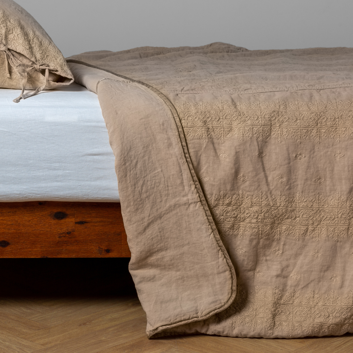 Honeycomb: White: embroidered bedspread with corner pulled back  showing the plain linen back. Shown with a white fitted sheet and matching sham, the bedframe is slightly visible  and the bed is against a white wall and on a medium wood floor. 