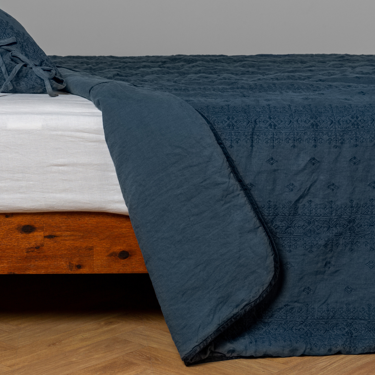 Midnight: White: embroidered bedspread with corner pulled back  showing the plain linen back. Shown with a white fitted sheet and matching sham, the bedframe is slightly visible  and the bed is against a white wall and on a medium wood floor. 