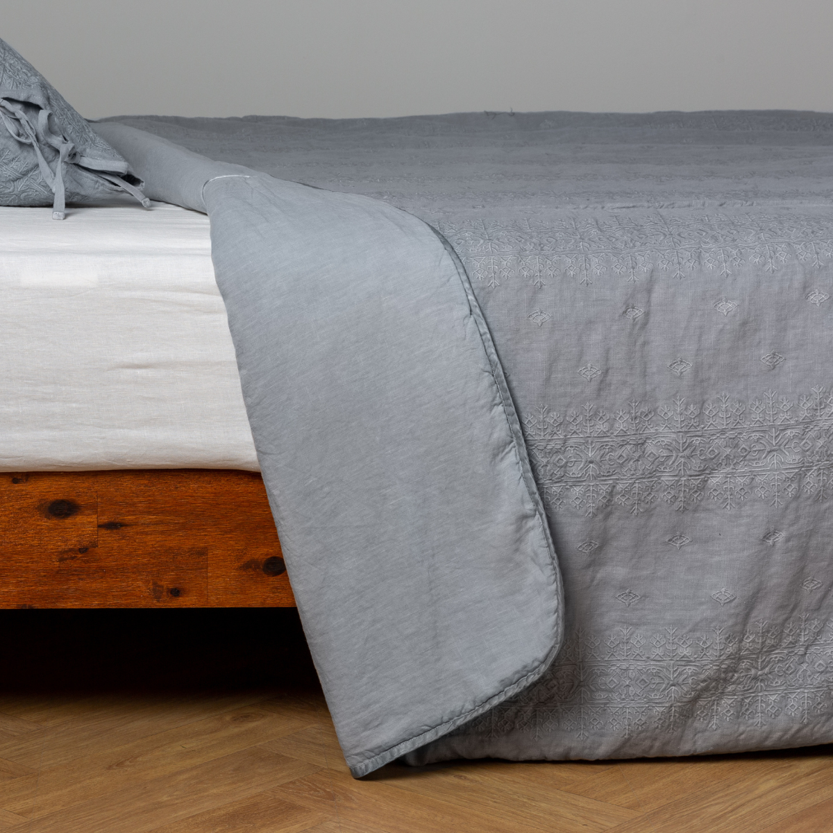 Mineral: White: embroidered bedspread with corner pulled back  showing the plain linen back. Shown with a white fitted sheet and matching sham, the bedframe is slightly visible  and the bed is against a white wall and on a medium wood floor. 