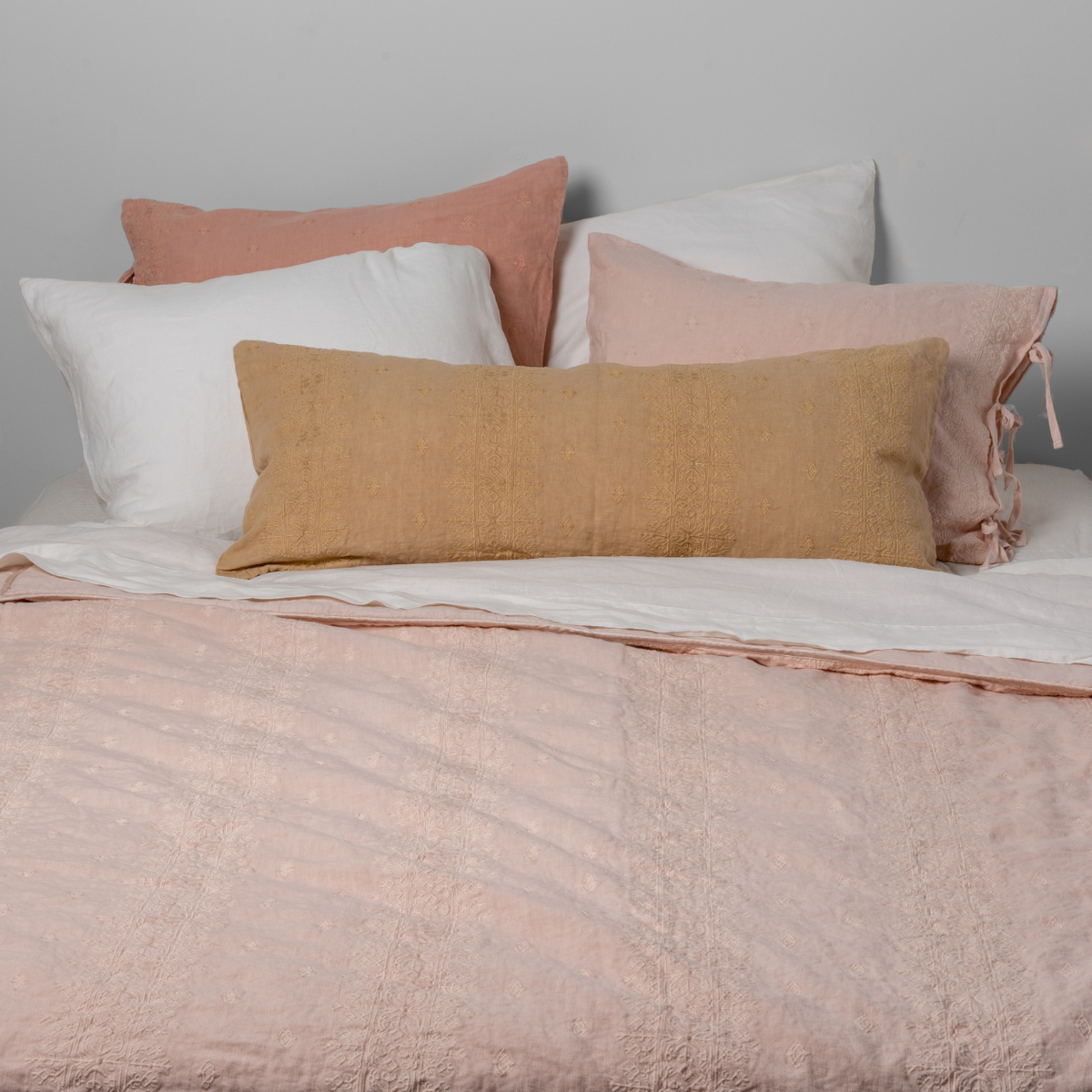 : Embroidered bedspread shown in pearl with white sheets and  sleping pillows, and warm toned shams and throw pillow. Shot from the end of the bed and the sheet is pulled back over the top of the  bedspread.