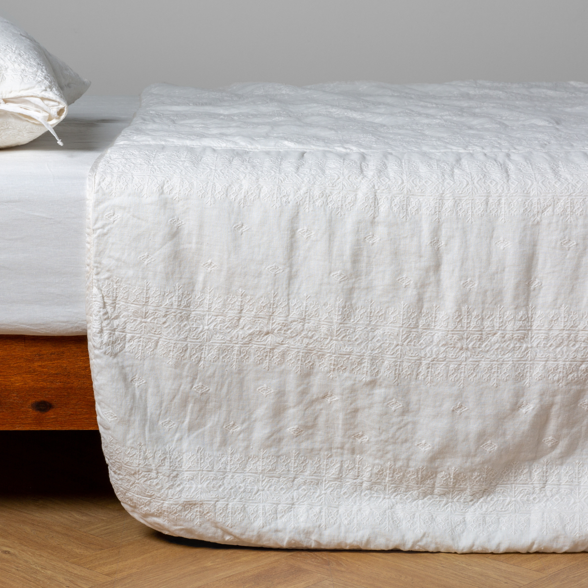 Winter White:  embroidered midweight linen bedspread with matching sham on a bed with a white fitted sheet. Shot from the side view, the bedframe is slightly visible  against a white background and medium wood floor. 