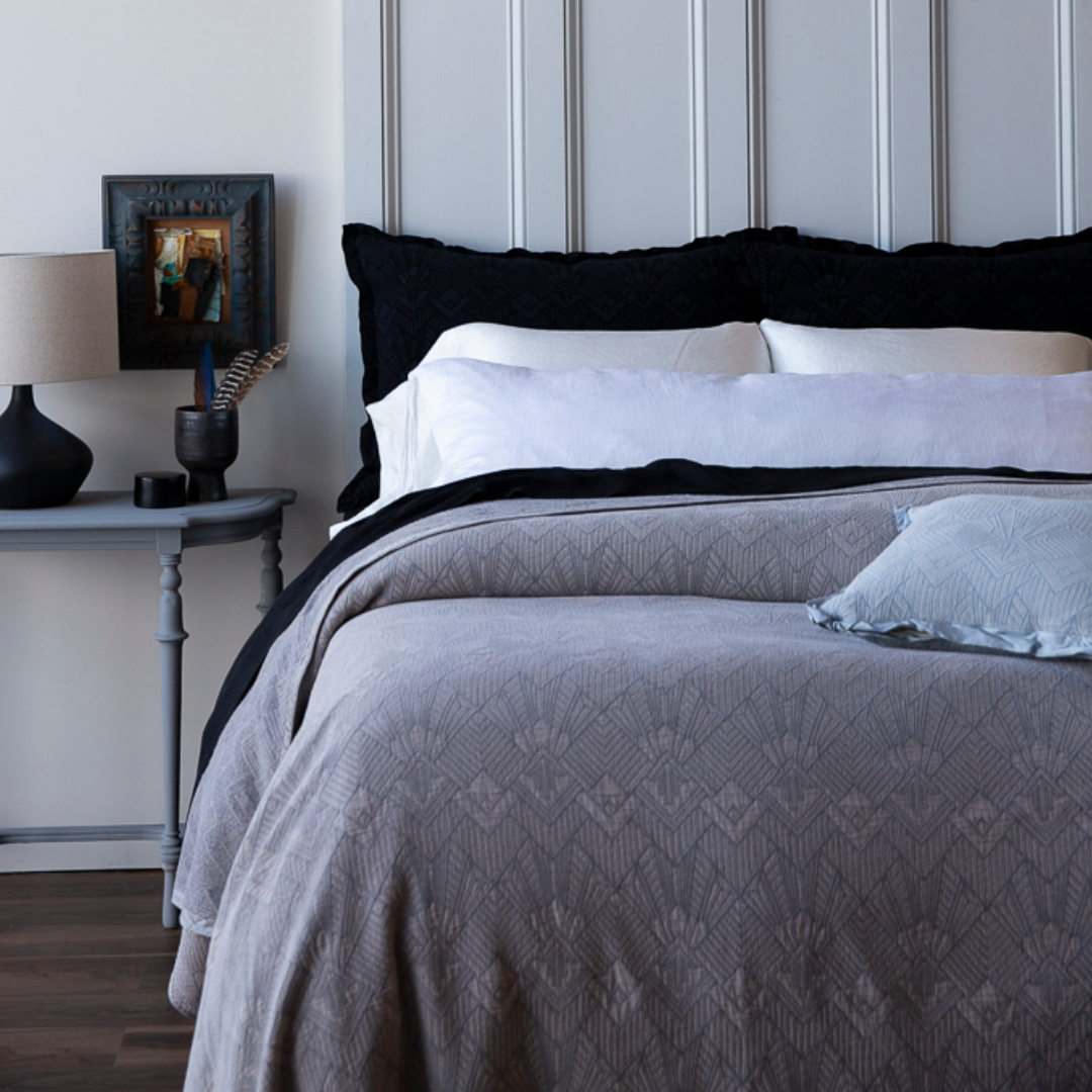 [allvariants]: Bedroom with a bed featuring gray bedding and black pillows, nightstand with lamp and decor.