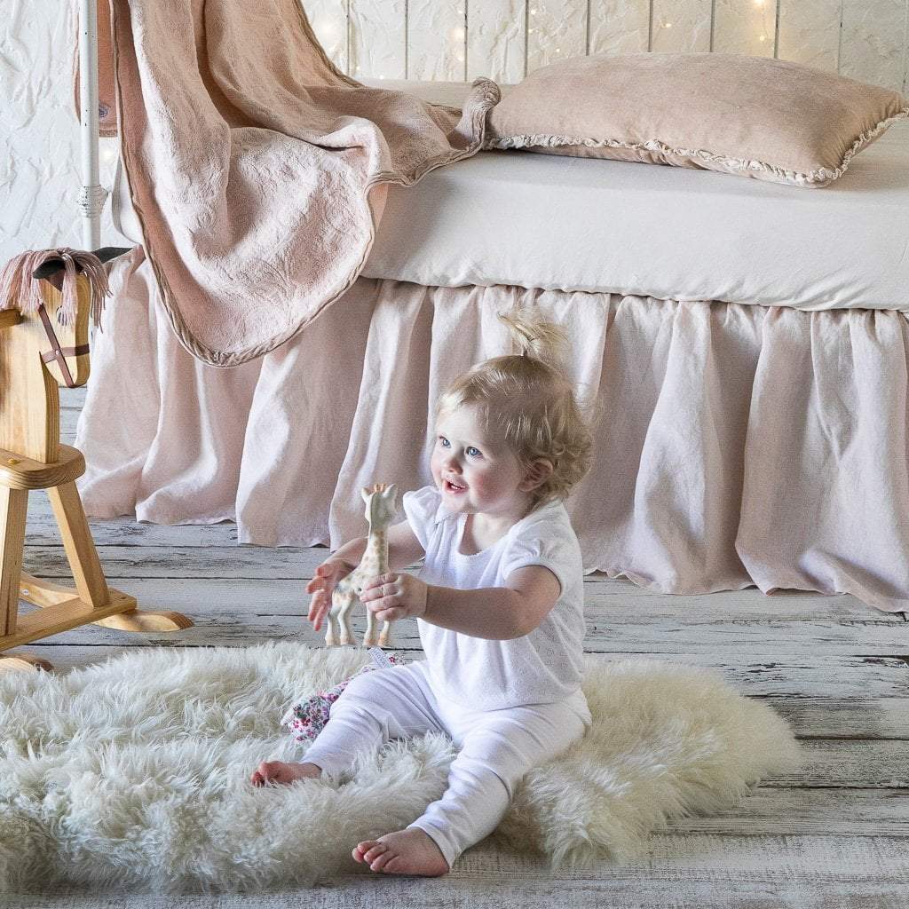 : Madera Luxe crib sheet in pearl with on-tone baby blanket, pillow, and crib skirt on a white iron crib. Side view with baby sitting and playing in the foreground.