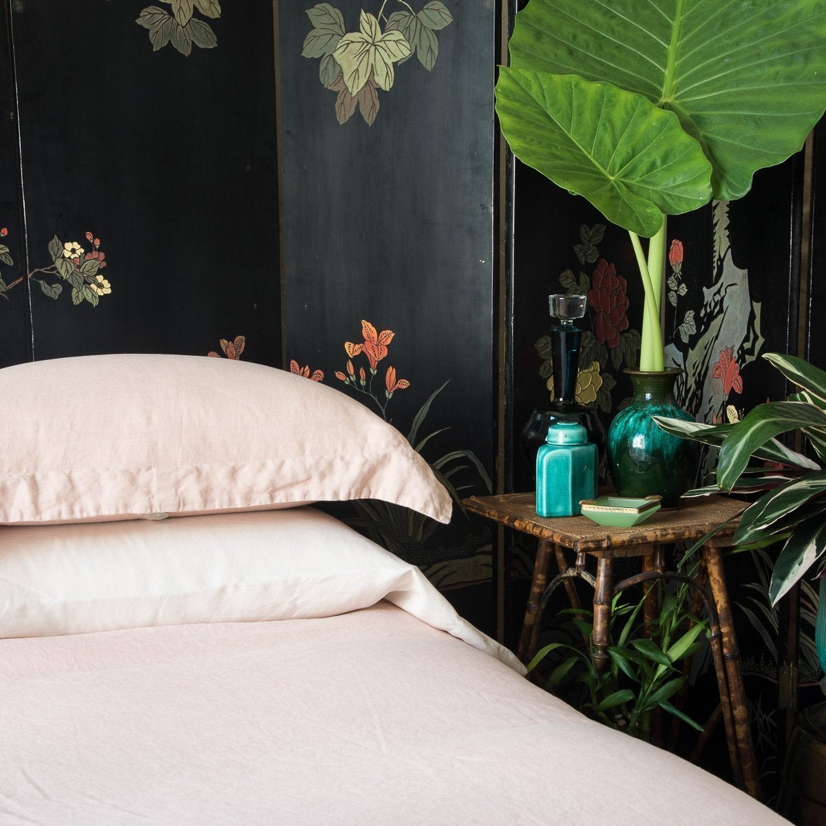 : Madera Luxe sleeping pillow laid flat under a linen sham, on a linen duvet cover, all in pearl, with a dark floral wall panel and tall greenery - cropped three-quarter angle.
