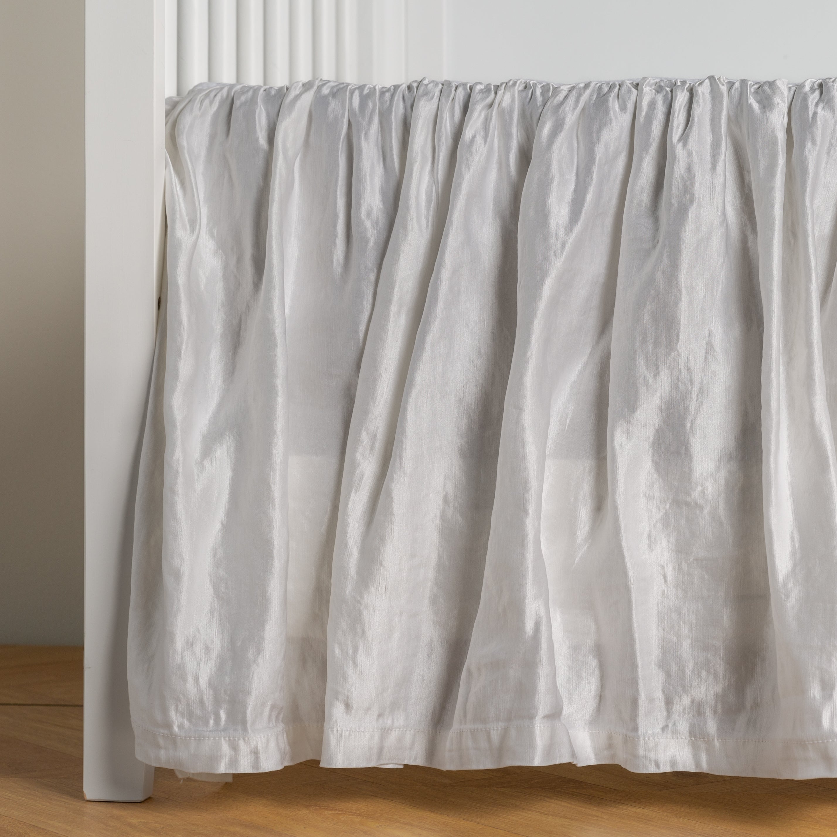 White: charmeuse crib skirt shown on a white crib against a white wall and medium wood floor. 
