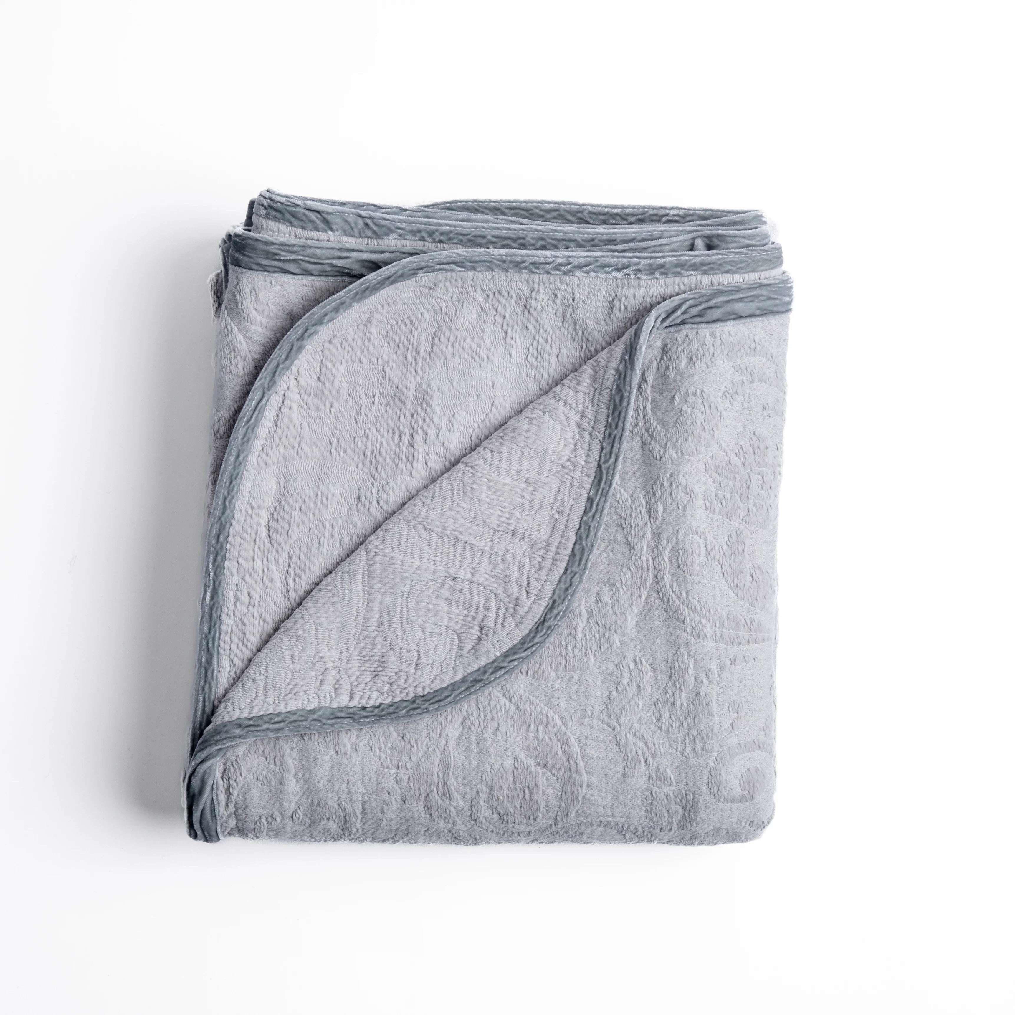 Sterling: folded organic cotton damask throw blanket shown with the corner folded down to show silk velvet trim detail — overhead against a plain white background. 