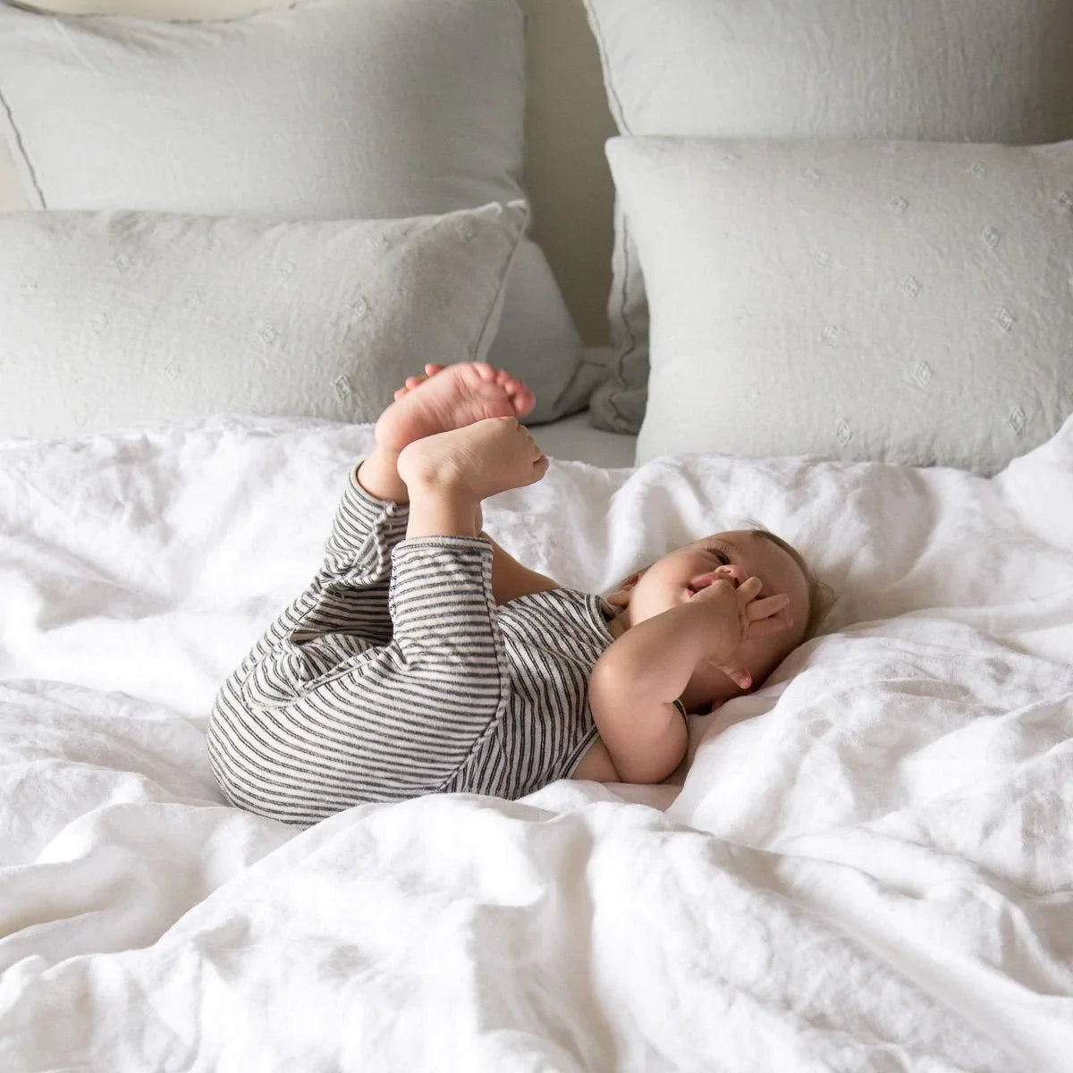 : Playful baby laying on rumpled Austin duvet cover with shams and pillows in the background.