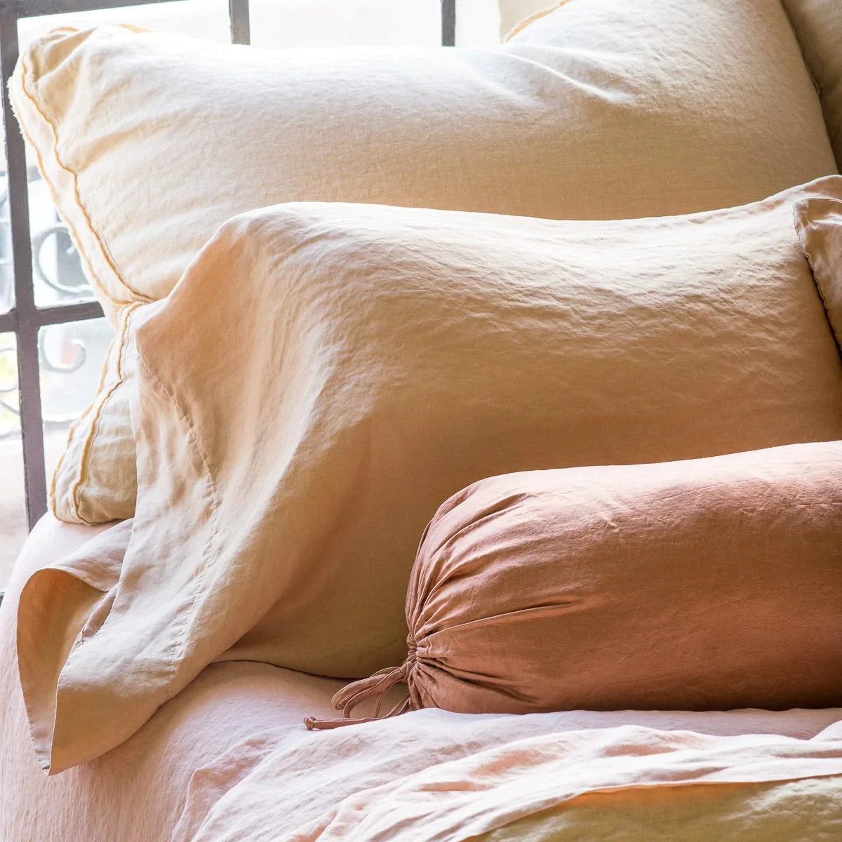 : Close up of Austin sham leaning against a black framed window, layered behind shimmery silk charmeuse pillows.