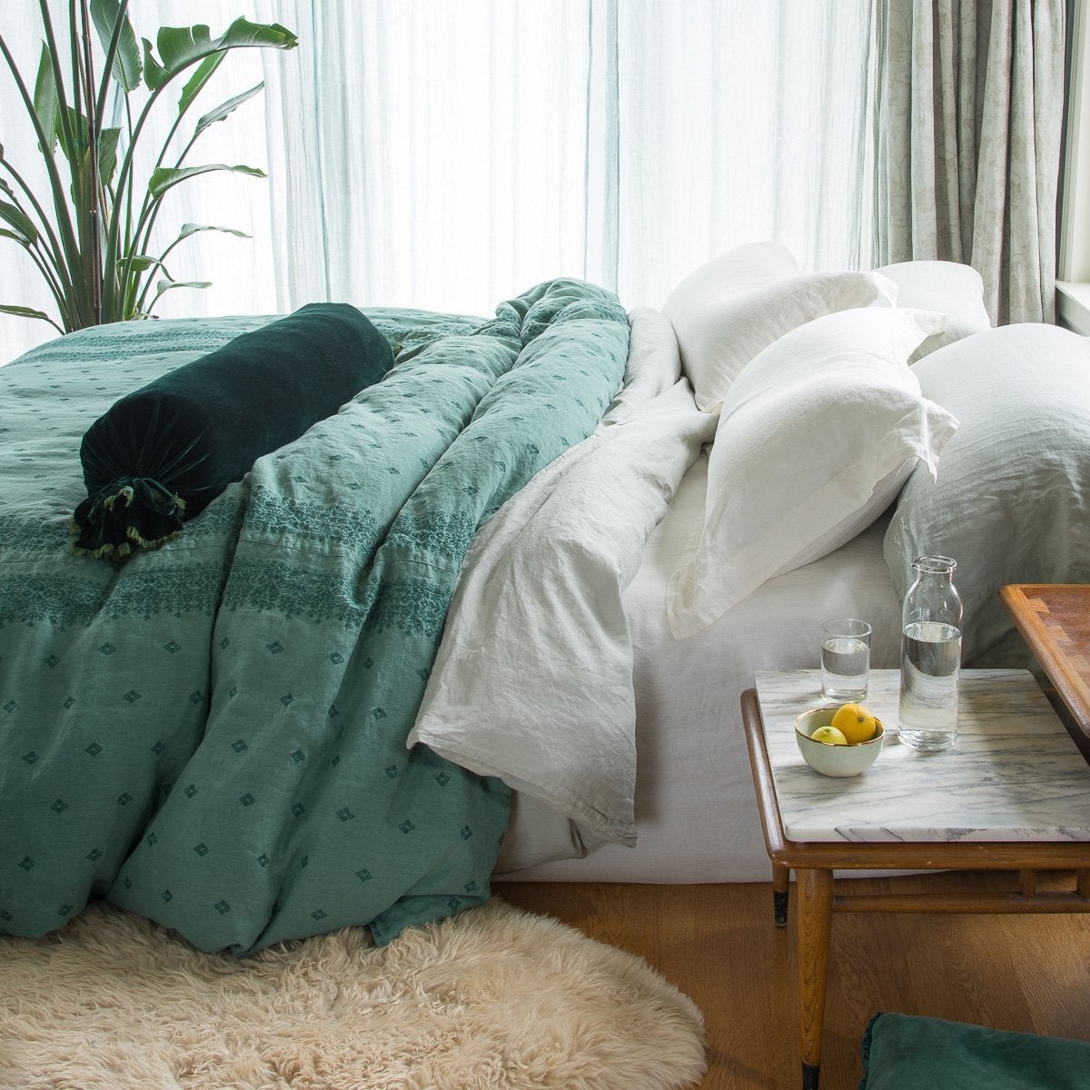 : Ines duvet cover in jade, lightly rumpled on a bed with linen sheets and pillows in soft white and green tones, complemented by warm wood in the foreground and sheer curtains in the background - side view.