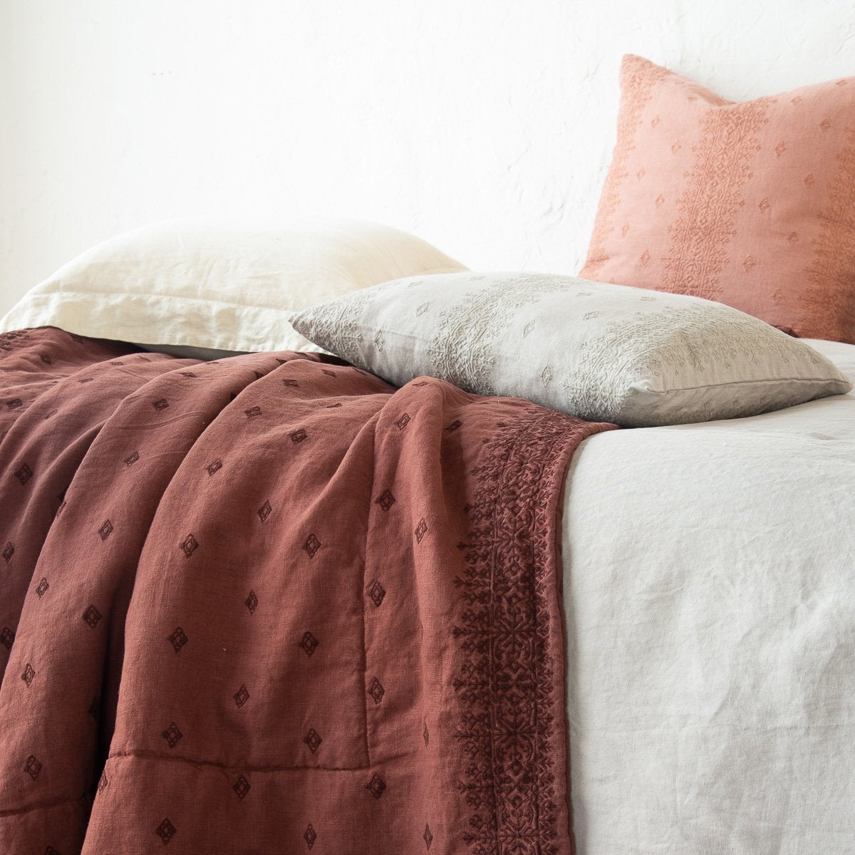 : Ines 24 by 24 pillow leaning upright against a textured white wall, on a bed with cream, soft blue, and brown accessory pieces - close-up side view.