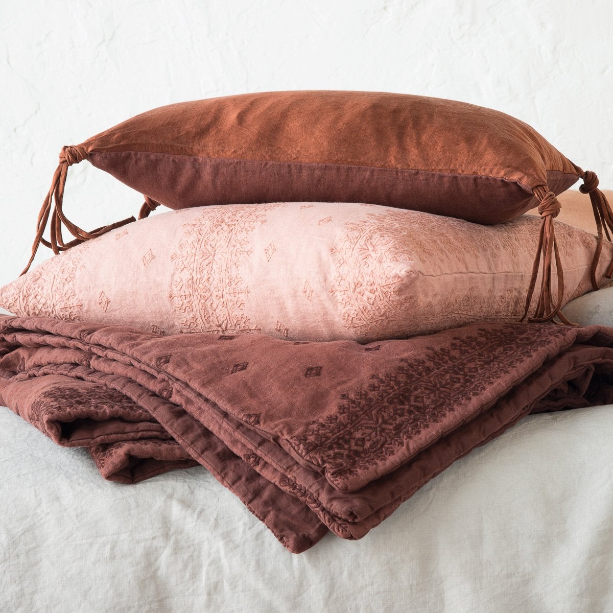 : Ines 24 by 24 pillow in rouge, stacked between a mahogany Ines folded throw blanket and a silk charmeuse accent pillow - side view.