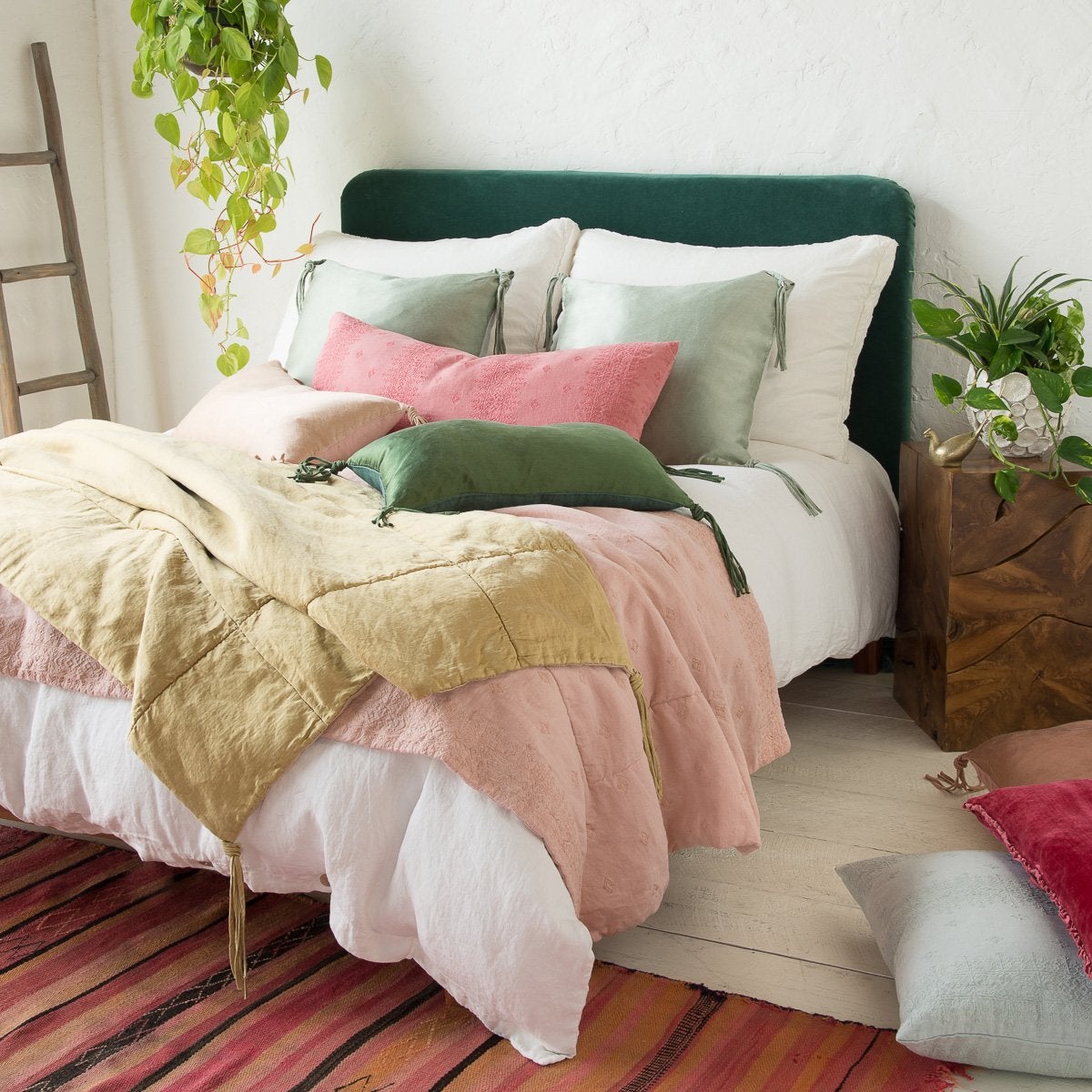 : Ines throw blanket in rouge, layered under green, pink, and gold tones on a white bed, surrounded by white walls, warm wood, colorful striped rug, and lush greenery - three-quarter angle view.