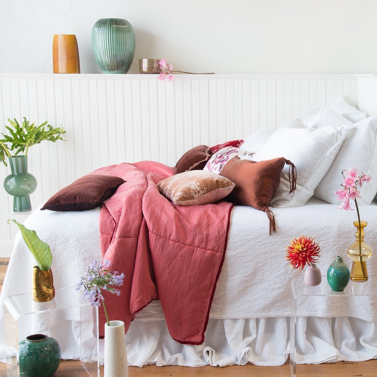 : Linen bed skirt in white, layered on an mostly white bed with rich pink and brown accents, and surrounded by colorful vases - side view.