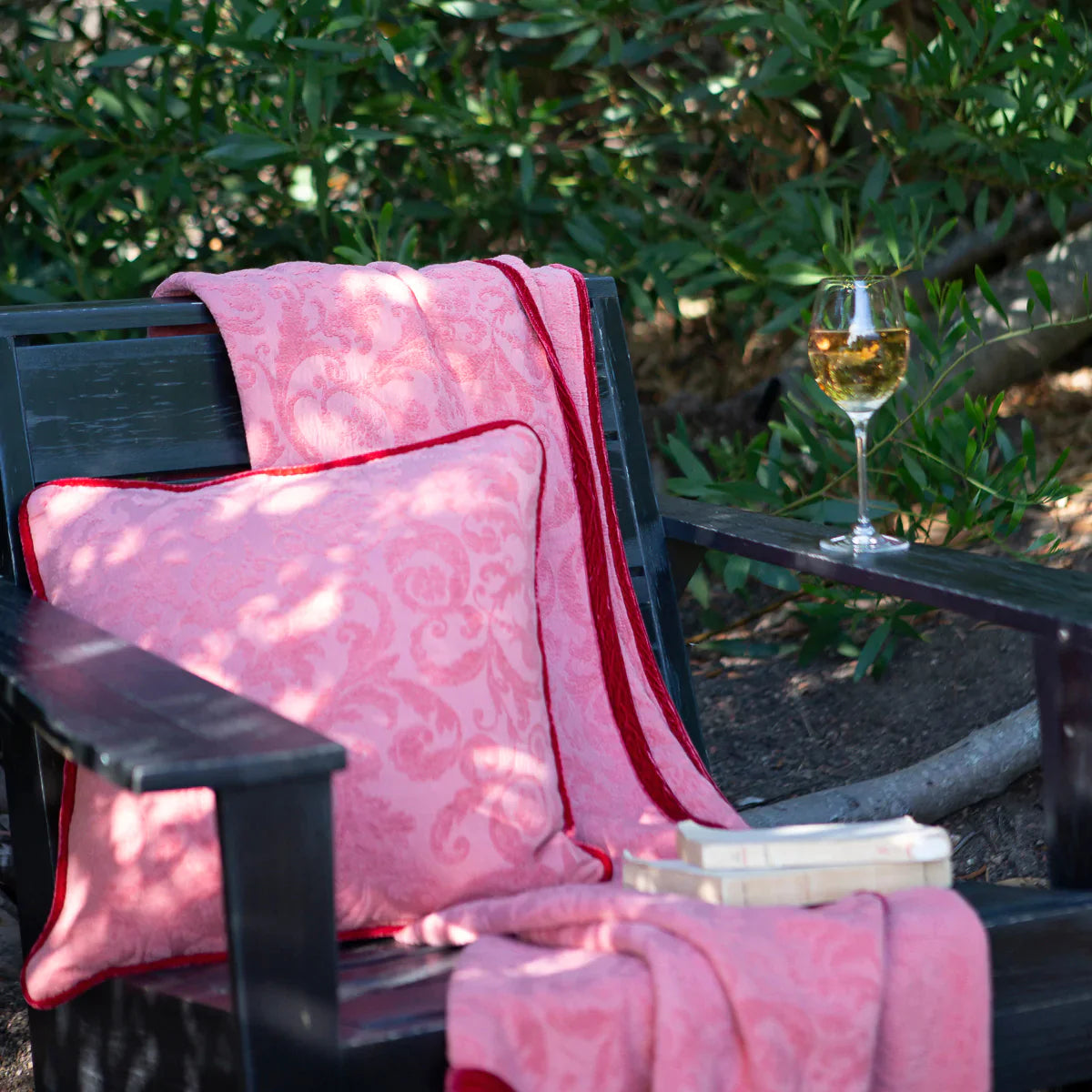 : matching organic cotton damask throw blanket and 18" square pillow in Poppy on an outdoor chair with a glass of white wine on the armrest and two paperback books on the seat. Greenery is visible behind the chair. 