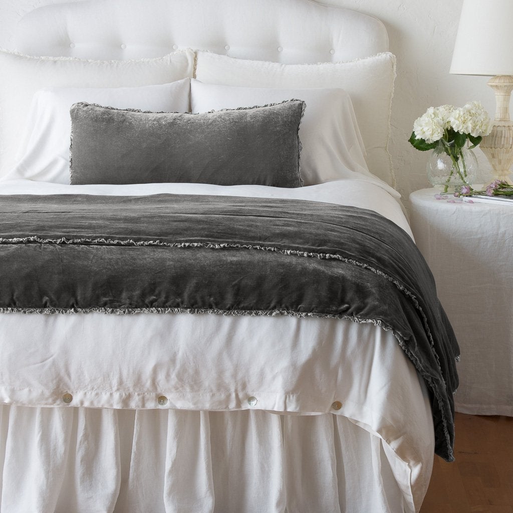 Fog: Silk velvet lumbar pillow with petite ruffle and throw blanket shown on an all white, neatly made bed - foot of bed angle.