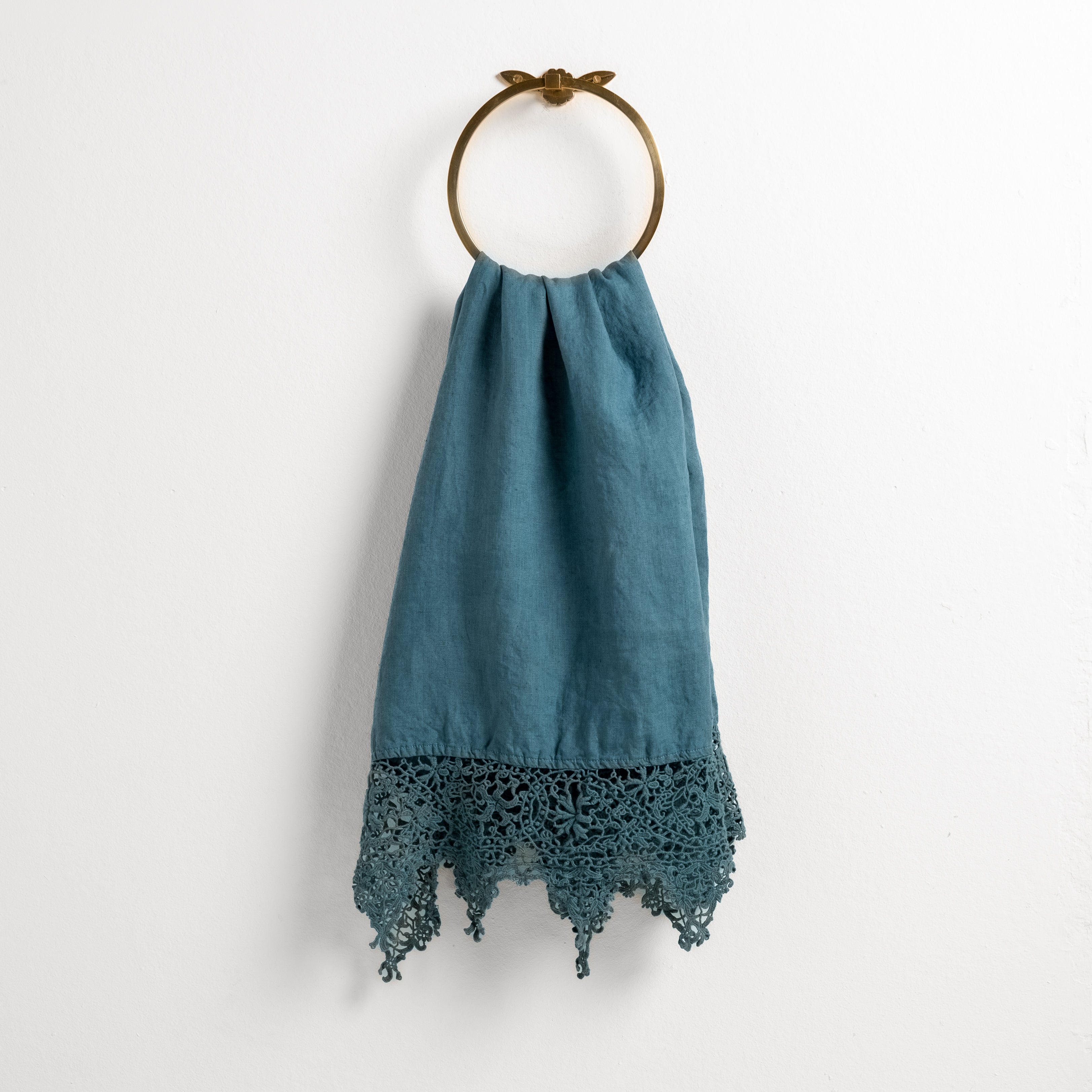 Cenote: Lace trimmed linen guest towel  draped through a decorative towel ring against a plain white background.