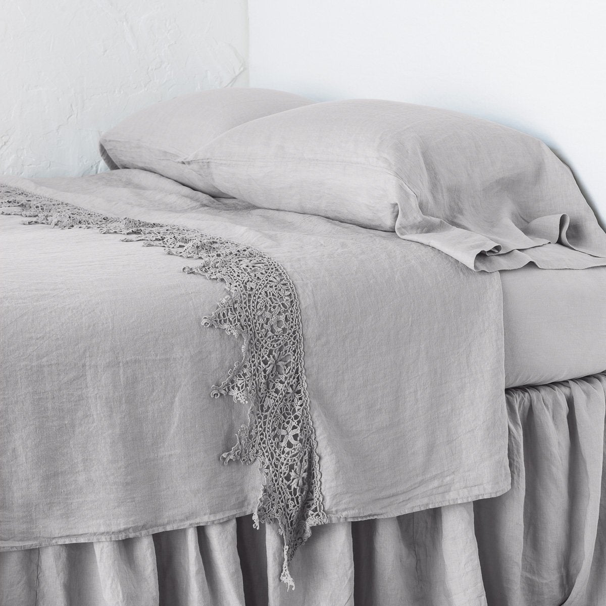 Fog: Lace trimmed linen flat sheet neattly folded back on a monochromatic linen bed - cropped three-quarter angle.
