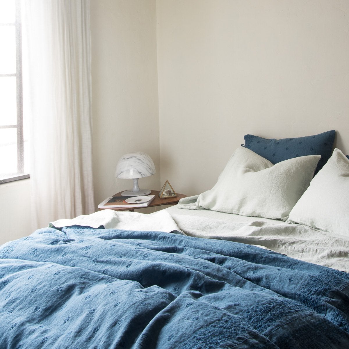: Midnight Ines duvet cover layered with soft green linen sheets, partially unmade with light toned walls and curtains, all bathed in natural light - cropped three-quarter angle.