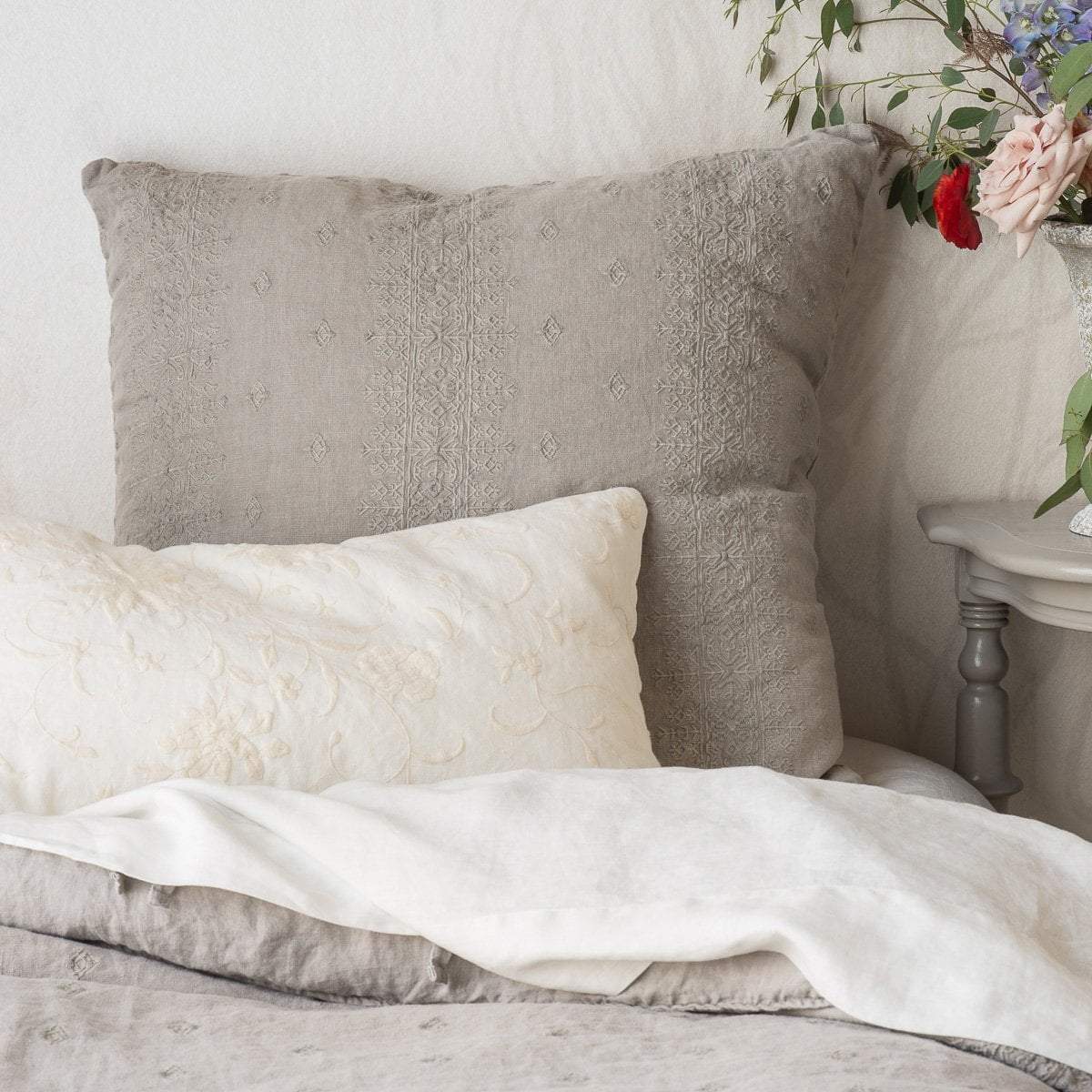 : Ines euro sham in fog, against a white wall, with cream tone sheets and throw pillow, next to a vase of colorful flowers - cropped head-on view.
