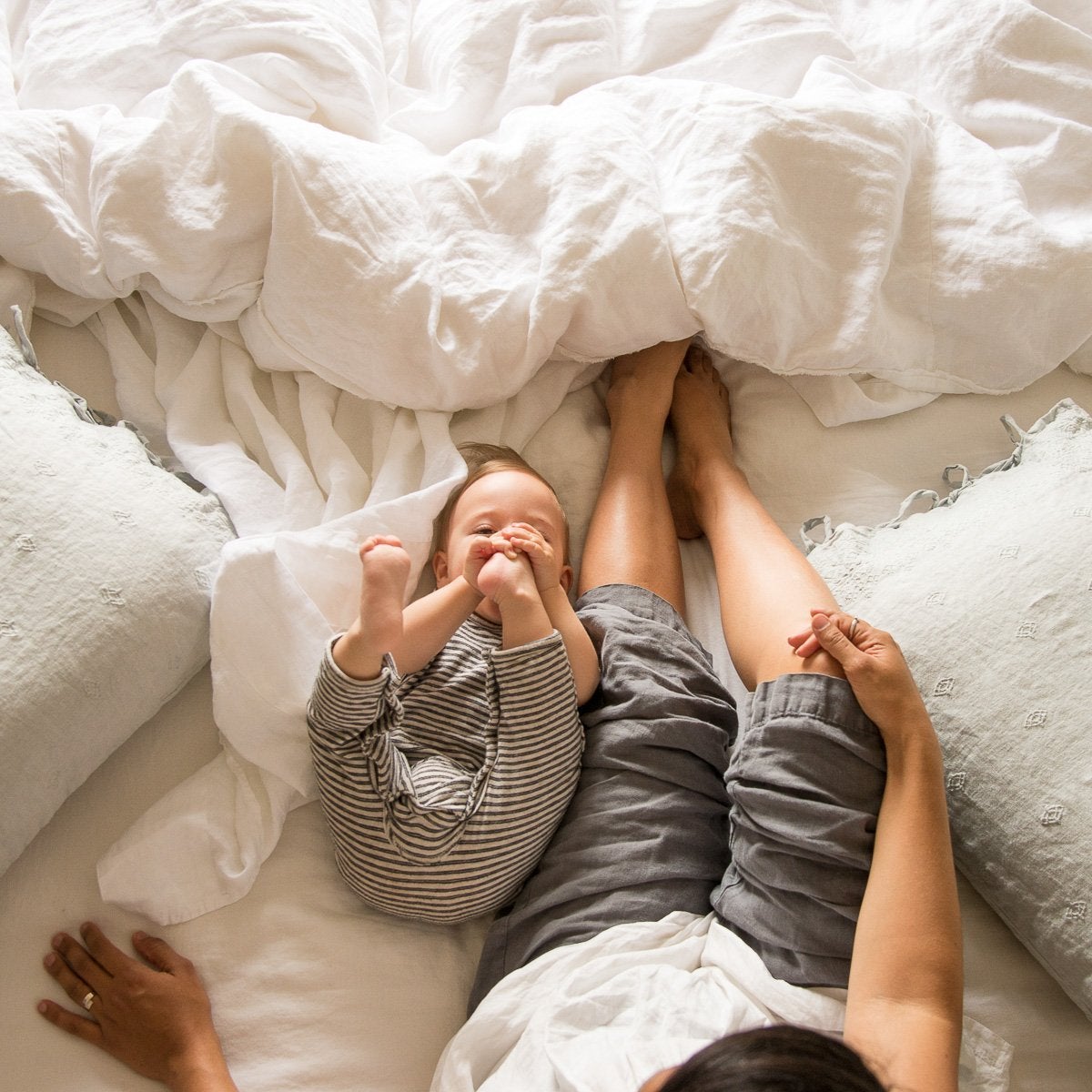 : Mother and baby relaxing on a white linen bed with Ines standard shams in eucalyptus on either side - overhead view.